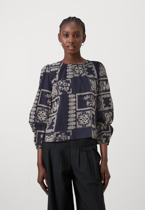 Marinebluse mit cremfarbenen, komplexen geometrischen und floralen Mustern, runder Ausschnitt und puffärmeligen Ärmel, kombiniert mit schwarzen, weit geschnittenen Hosen.
