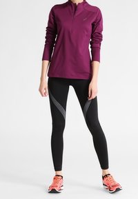 Långärmad, lila sporttröja med halv dragkedja, matchad med svarta leggings med ett grått zigzag-mönster och orange löparskor.