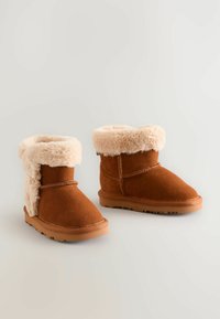 Brune ruskind ankelstøvler med en fluffy lys tan pelsfoering og kant. Sålene er af gummi med et tekstureret grebsmønster.