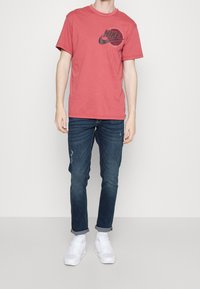 Camiseta de algodón roja con un logo negro de Nike, combinada con jeans azul oscuro y zapatillas blancas, con un ligero desgastes en los jeans.