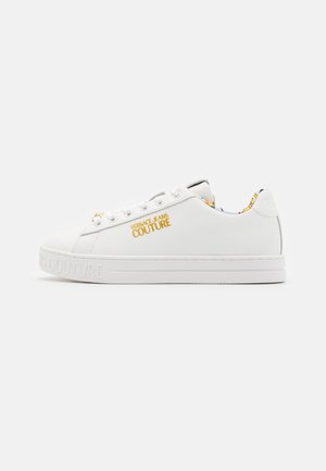 Basket blanche en matériau synthétique lisse, avec un bout rond, une semelle blanche et une marque dorée "VERSACE JEANS COUTURE" sur le côté.