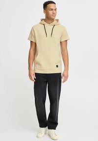 Beige hættebluse med korte ærmer, struktureret stof, forlommek, sorte snore, parret med sorte jeans og hvide lavtop sneakers.