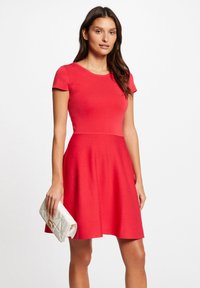 Rotes kurzärmliges Kleid mit tailliertem Oberteil und ausgestellt Rock, aus strukturierter Stoff gefertigt. Kombiniert mit einer weißen, gesteppten Clutch mit goldenen Beschlägen.