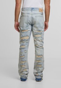 Šviesiai mėlyni džinsai, pasižymintys laisvu siluetu, suplėšytomis vietomis, suplūkusiais kraštais ir šviesiai rudos spalvos prekės ženklo etiketėmis ant užpakalinės juostos.