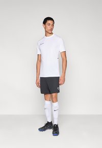 Nike sportöltözet: fehér rövid ujjú felső, fekete rövidnadrág, fehér térd fölötti zokni és kék részletekkel díszített fekete stoplis cipő.