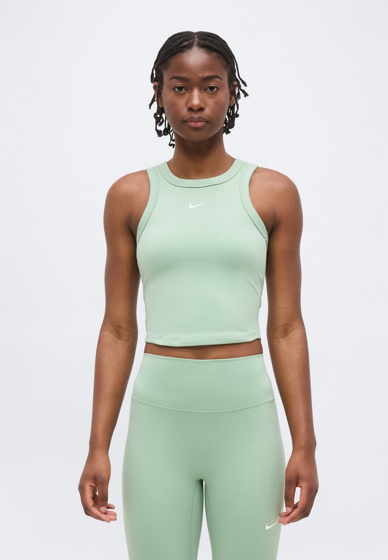 Νέα γυναίκα με πλεξούδες που φοράει ανοιχτό πράσινο αμάνικο crop top και ταίριασμα ψηλόμεσων κολάν, και τα δύο με μικρά λευκά λογότυπα Nike.