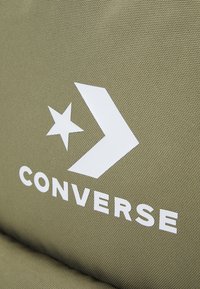 Logo bianco di Converse con stella e chevron su sfondo di tessuto verde oliva texturizzato.