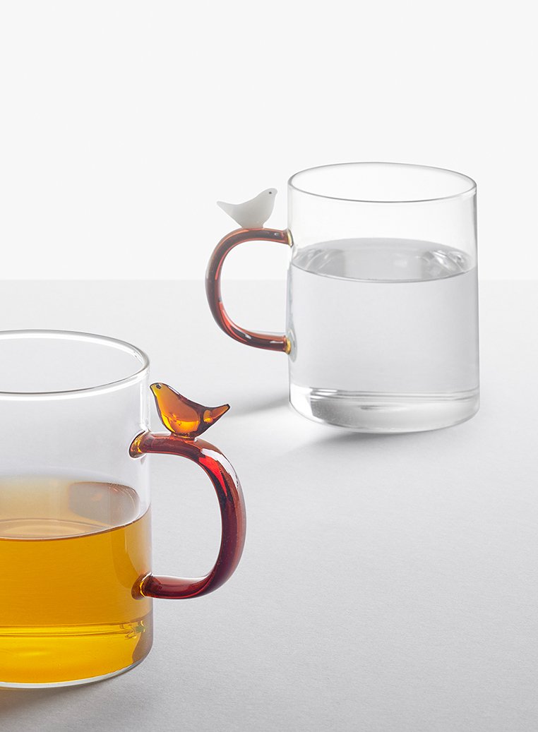 Dwa przejrzyste szklane kubki, jeden napełniony bursztynowym płynem, drugi wodą, każdy z zaokrąglonym brązowym uchwytem zwieńczonym małą ozdobną figurką ptaka.