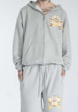 Person iført en lysegrå hættetrøje med lynlås og matchende træningsbukser, begge med gule og hvide grafiske logoer på bryst og lår.