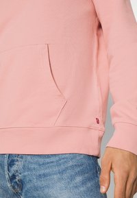Sweat-shirt rose avec poche avant, texture douce et poignets côtelés. Présente une étiquette logo rouge sur l'ourlet. Porté avec un jean bleu clair.