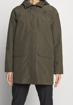 Persona con chubasquero verde oliva con capucha, cierre de botones en la parte delantera y bolsillos con solapas, combinado con pantalones negros, de pie frente a un fondo liso.