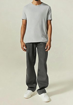 Homme portant un t-shirt gris clair à manches courtes, un pantalon noir ample et des baskets blanches, debout devant un fond clair uni.