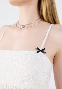 Bílé krajkové šaty s tenkými ramínky; na jednom ramínku je malá černá mašlička; strukturovaná tkanina přidává hloubku designu.