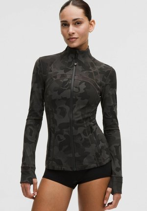 Kobieta w dopasowanej czarno-szarej kurtce moro na suwak z otworami na kciuki i czarnych szortach, pozująca na jasnym tle.