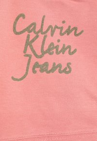 Prašný ružový textil s nápisom "Calvin Klein Jeans" v kontrastnej olivovo zelenej farbe. Materiál vyzerá mäkký s hladkou textúrou.