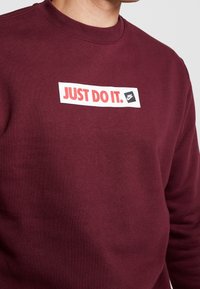 Mörkröd sweatshirt med rund halsringning och ribbade muddar, med en vit rektangulär patch med röd och svart text "JUST DO IT." och Nike-logotyp.