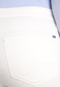 Jeans bianchi con una texture liscia, caratterizzati da una tasca posteriore con cuciture sottili e un piccolo tag argentato del marchio vicino alla vita.