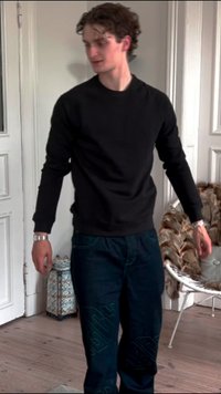 Schwarzes Sweatshirt mit langen Ärmeln, kombiniert mit blauen Jeans aus Denim mit hellblauen Akzenten. Innenraum mit hellem Holzfußboden.