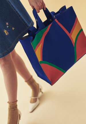 Femeie într-o fustă din denim care ține o geantă mare albastră cu modele abstracte colorate, purtând șosete bej și pantofi cu toc albi.