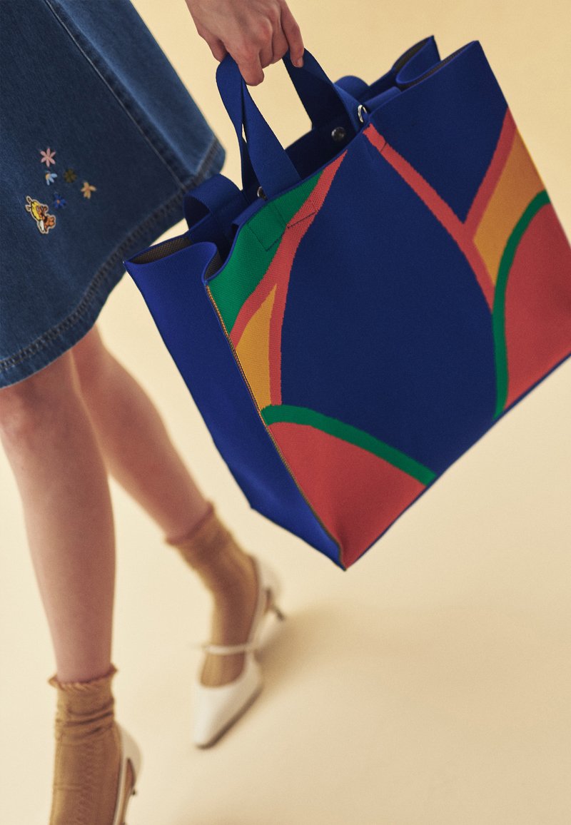 Femme en jupe en denim tenant un grand sac cabas bleu avec des motifs abstraits colorés, portant des chaussettes beiges et des talons blancs.