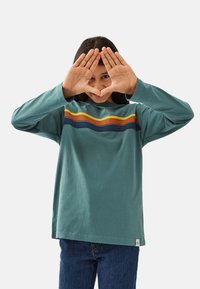 Langärmliges türkisfarbenes T-Shirt mit mehrfarbigen, horizontalen Streifen in Orange, Blau und Gelb über der Brust, getragen mit Jeans.