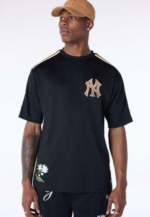 Hombre con camiseta negra con logo beige de Nueva York, gorra color beige y pantalones cortos negros, de pie con la cabeza girada ligeramente hacia la izquierda contra fondo blanco.