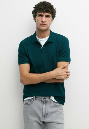 Ung mand med krøllet hår iført en mørkegrøn polo shirt over en hvid t-shirt og lysegrå jeans, står med armene krydset.