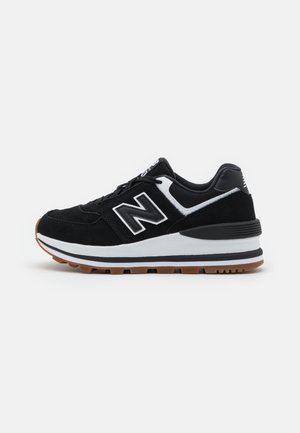 Zwart-witte New Balance sneaker met bruine zool, vetersluiting, bovenwerk van mesh en suède, zijaanzicht op witte achtergrond.