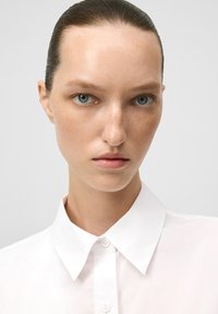 Giovane donna con occhi azzurri, pelle naturale e capelli pettinati all'indietro che indossa una camicia bianca con bottoni, su uno sfondo chiaro e uniforme.