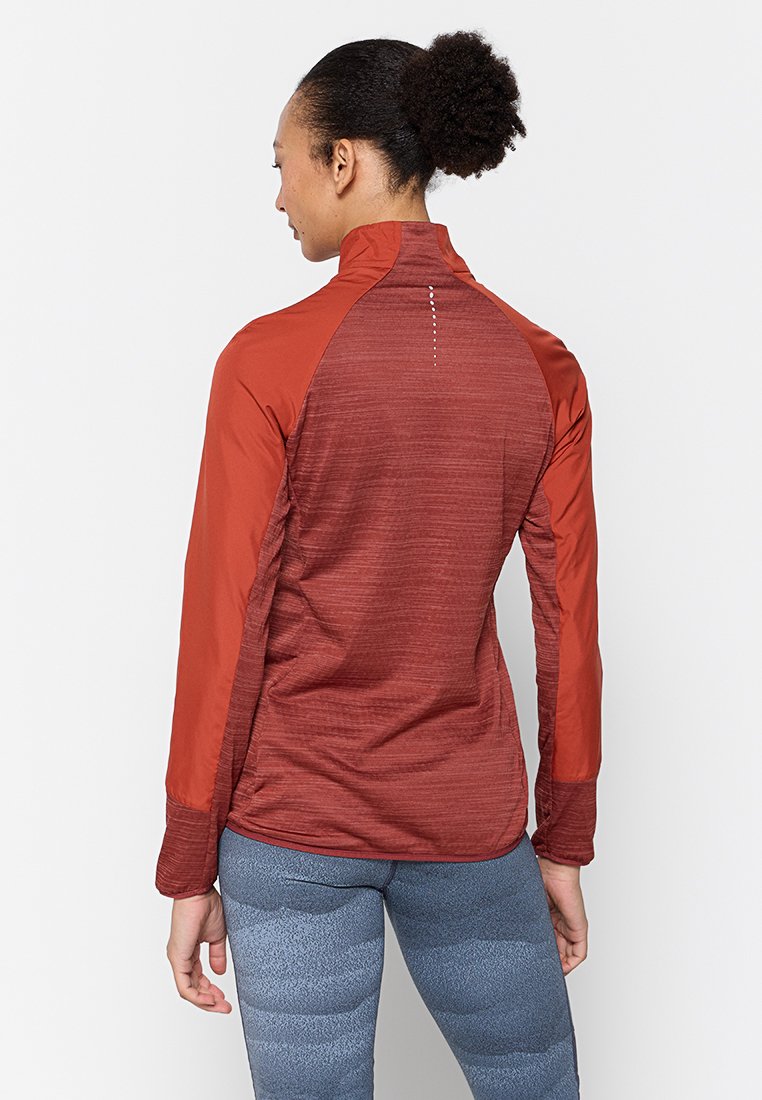 Frau mit lockigem Haar in einem Pferdeschwanz, die eine rostfarbene langärmlige Sportjacke und blaue gemusterte Leggings trägt, von hinten gezeigt.