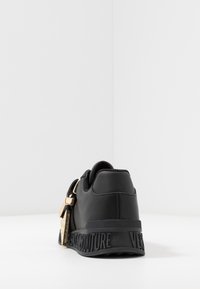 Baskets noires avec une texture en cuir lisse, détails en métal doré et marquage en relief sur la semelle en caoutchouc. Design bas avec une forme arrondie.