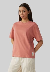 Różowa bawełniana koszulka z luźnym fasonem, krótkimi rękawami, okrągłym dekoltem i małą ozdobną naszywką na dolnym brzegu.