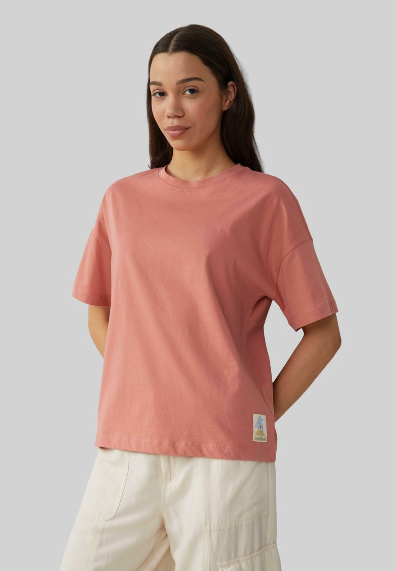 Różowa bawełniana koszulka z luźnym fasonem, krótkimi rękawami, okrągłym dekoltem i małą ozdobną naszywką na dolnym brzegu.