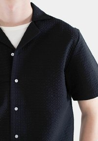Kortermet, svart skjorte med et teksturert mønster, knappelukking foran, og en klassisk krage. Stoffet ser vevd ut og har en lett glans.