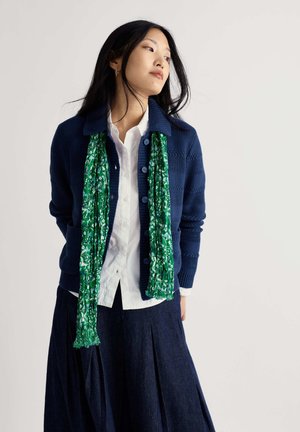 Femme aux longs cheveux noirs portant un gilet bleu marine, une chemise blanche, une écharpe verte à motifs et une jupe plissée foncée, regardant sur le côté.