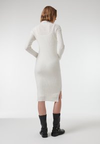 Lang wit gebreid jurk met ribbelstructuur, voorzien van een hoge kraag, een getailleerde vorm en een zijsplit in de zoom. Gemaakt met zwarte laarzen tot de knie.
