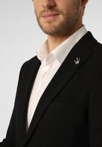 Schwarzes Blazer mit zartem Schimmer, darunter ein weißes Hemd und eine silberne Krone als Reversnadel. Strukturierte Passform mit Reverskragen.