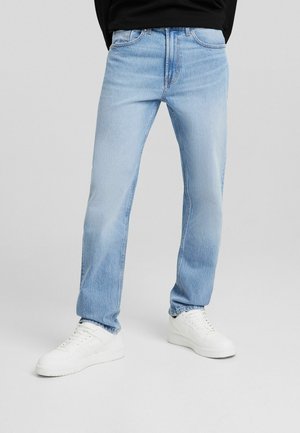 Man draagt lichtblauwe rechte spijkerbroek en witte sneakers, staand tegen een effen lichtgrijze achtergrond.