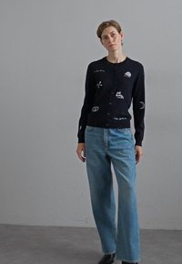 Marinblå stickad cardigan med broderade mönster, med knappar framtill. Kombination med ljusblå vida jeans och svarta skor.