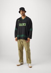 Čierne dlhé rukávy s dresom so zeleným textom "CELTICS" a pruhmi, v kombinácii s khaki nohavicami so bočnými patentkami a bielymi teniskami.