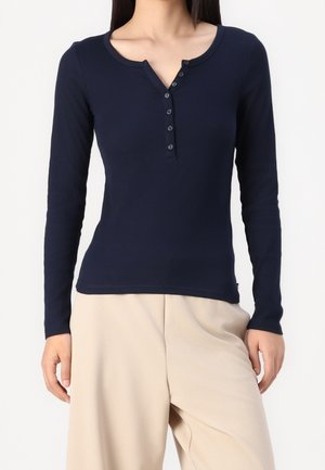 Vrouw die een marineblauw lang mouw henley-shirt met zes knopen draagt, gecombineerd met beige wijde broekspijpen tegen een effen achtergrond.