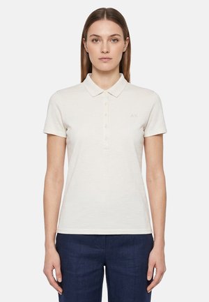 Donna in piedi rivolta in avanti, indossa una polo a maniche corte, semplice, color avorio chiaro e pantaloni blu scuro, su sfondo bianco.