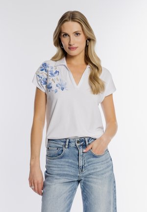 Blondynka w białej koszulce z krótkim rękawem i dekoltem w serek, ozdobionej niebieskim kwiatowym haftem na ramieniu, oraz w jasnoniebieskich dżinsach, stojąca z ręką w kieszeni.