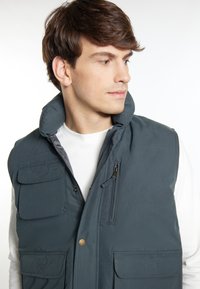Dunkelgraue Herren-Pufferweste mit hohem Kragen, mehreren vorderen Taschen und Reißverschlüssen; glatte Textur mit gesteppten Details.