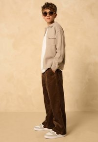 Helles beige Strickpullover mit Kragen, kombiniert mit braunen Cordhosen und weißen Sneakers mit grauen und roten Akzenten. Model trägt eine dunkle Sonnenbrille.