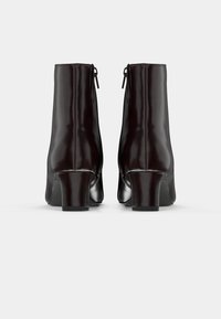 Pár tmavě hnědých kožených kotníkových bot s blokovými podpatky, zobrazených zezadu na jednoduchém světlém pozadí.