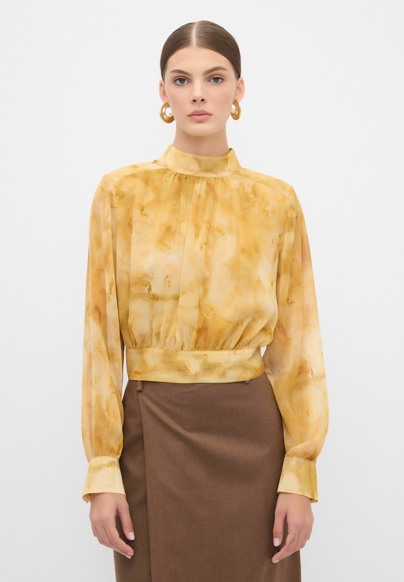 Gelbe, gemusterte Bluse mit langen Ärmeln, hohem Kragen und gerafftem Detail an der Taille, kombiniert mit einem engen braunen Rock.
