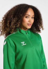 Groene sportjack met een kwart ritssluiting, voorzien van witte accenten op de schouders en een logo op de borst; gladde stof en getailleerd ontwerp.