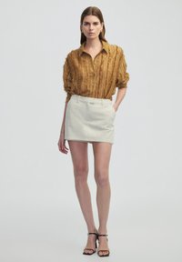Brown textured shirt with a loose fit, paired with a light gray mini skirt. Model poses with black heeled sandals, showcasing clean lines and simplicity.
