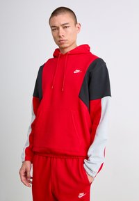 Tânăr îmbrăcat într-un hanorac Nike roșu, negru și alb, împreună cu pantaloni de trening Nike roșii asortați, stând în fața unui fundal deschis.